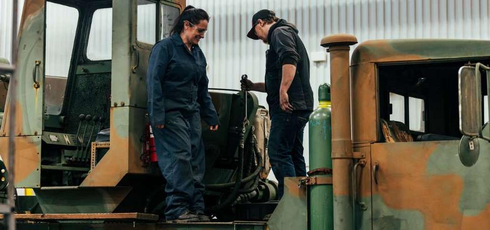 A male and female student standing on a military painted truck laughing while one is holding a a wrench about to tighten a hose